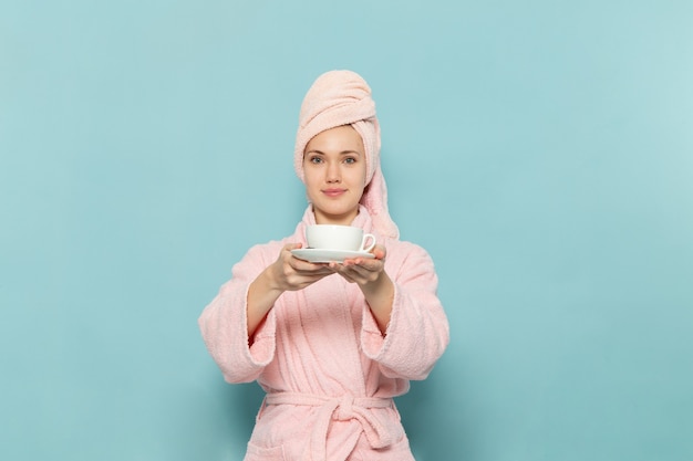 Hembra joven en bata de baño rosa después de la ducha bebiendo café sonriendo sobre azul