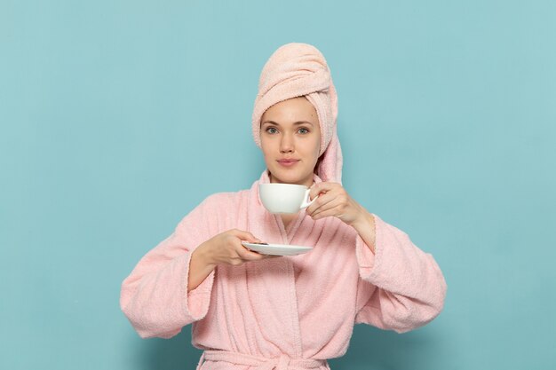 Hembra joven en bata de baño rosa después de la ducha bebiendo café sonriendo sobre azul