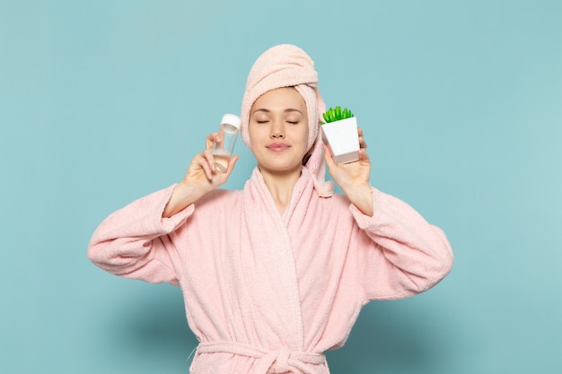 Hembra joven en bata de baño rosa después de la ducha sosteniendo spray y planta verde sobre azul