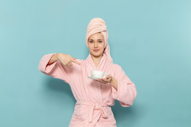 Hembra joven en bata de baño rosa después de la ducha sosteniendo una taza de café blanco en el escritorio azul