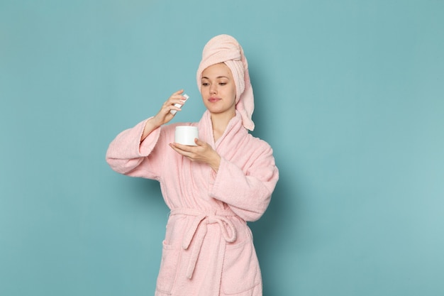 Hembra joven en bata de baño rosa después de la ducha sosteniendo y usando crema en azul