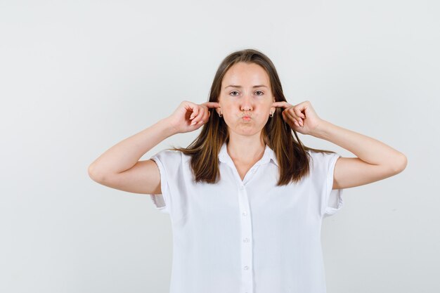 Hembra joven en blusa blanca taponando las orejas con los dedos y mirando saciado