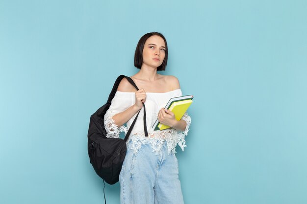 Hembra joven en camisa blanca, blue jeans y bolso negro sosteniendo cuadernos pensando en azul