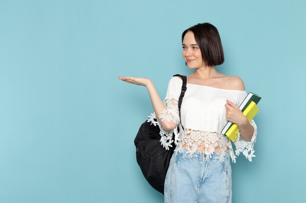 Hembra joven en camisa blanca, blue jeans y bolso negro sosteniendo cuadernos sonriendo sobre azul