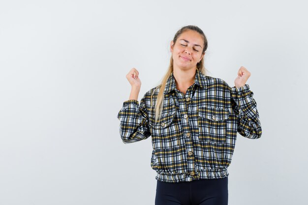 Hembra joven en camisa a cuadros que muestra el gesto del ganador y mirando alegre, vista frontal.