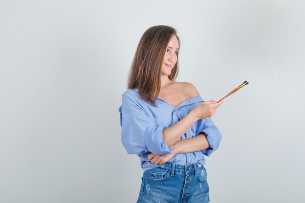 Hembra joven en camisa, pantalones cortos sosteniendo pinceles y mirando alegre