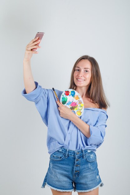 Hembra joven en camisa, pantalones cortos tomando selfie con herramientas de pintura