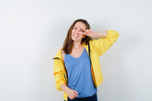 Hembra joven en camiseta, chaqueta que muestra el signo V en el ojo y parece confiada, vista frontal.