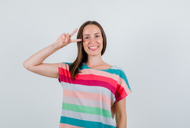 Hembra joven en camiseta mostrando v-sign y mirando feliz, vista frontal.