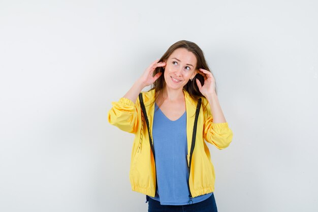 Hembra joven cogidos de la mano cerca de las orejas en camiseta, chaqueta y aspecto soñador. vista frontal.