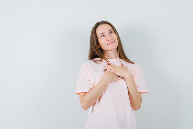 Hembra joven cogidos de la mano en el pecho en camiseta rosa y luciendo esperanzados. vista frontal.