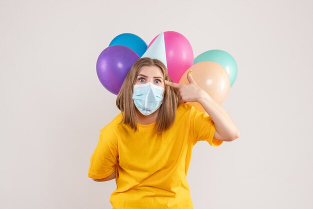 Hembra joven escondiendo globos de colores detrás de su espalda en máscara en blanco