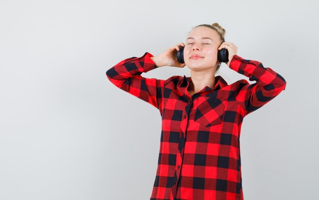 Hembra joven escuchando música con auriculares en camisa a cuadros y mirando encantada. vista frontal.