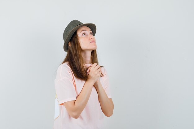 Hembra joven estrechando las manos en gesto de oración en camiseta rosa, sombrero y mirando esperanzado. vista frontal.