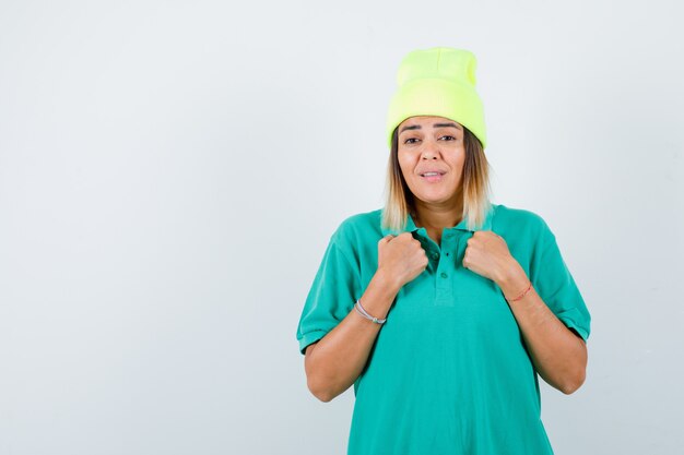 Hembra joven manteniendo los puños cerrados en el pecho en camiseta de polo, gorro y mirando gruñón, vista frontal.