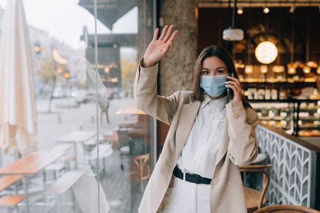 Hembra joven en mascarilla en café durante la cuarentena. Mujer de negocios trabajando en cuarentena. Mujer habla por teléfono. COVID-19