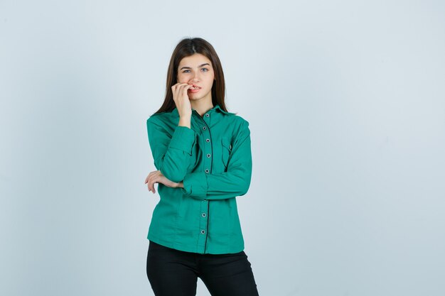 Hembra joven mordiendo las uñas en camisa verde, pantalones y aspecto preocupado. vista frontal.