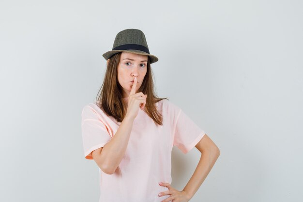 Hembra joven mostrando gesto de silencio en camiseta rosa, sombrero y mirando con cuidado, vista frontal.