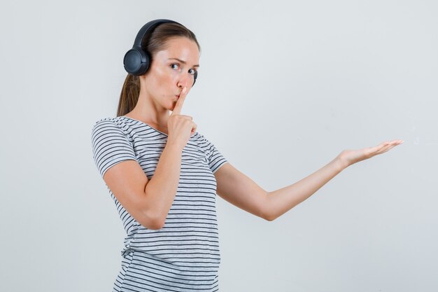 Hembra joven mostrando gesto de silencio con la palma de la mano en camiseta, auriculares, vista frontal.