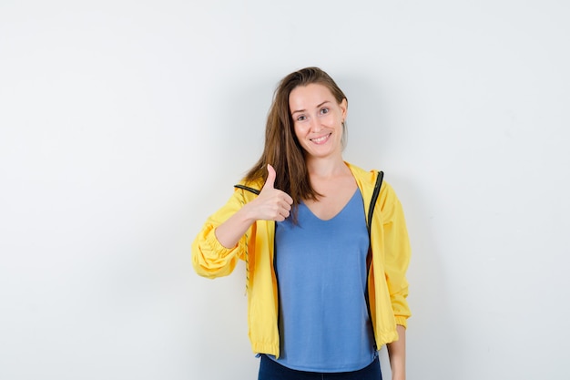 Hembra joven mostrando el pulgar hacia arriba en camiseta, chaqueta y mirando feliz. vista frontal.