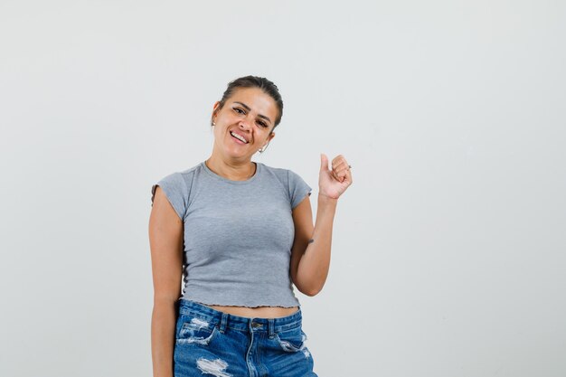 Hembra joven mostrando el pulgar hacia arriba en camiseta, pantalones cortos y mirando feliz