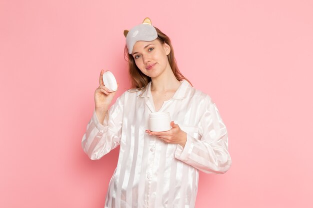 Hembra joven en pijama y antifaz para dormir posando con una sonrisa sosteniendo crema en rosa