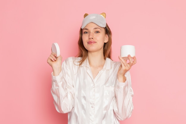 Hembra joven en pijama y antifaz para dormir sosteniendo crema y sonriendo en rosa