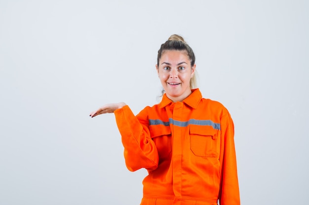 Hembra joven que extiende a un lado su mano con la palma abierta en uniforme de trabajador y parece complacida. vista frontal.