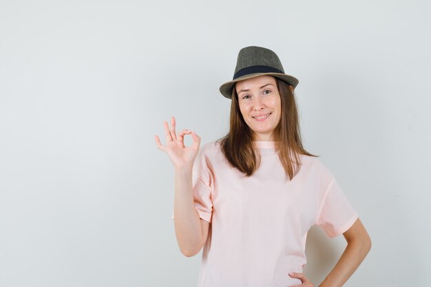 Hembra joven que muestra un gesto aceptable en camiseta rosa, sombrero y que parece seguro. vista frontal.