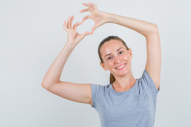 Hembra joven que muestra el gesto del corazón en camiseta gris y parece alegre. vista frontal.