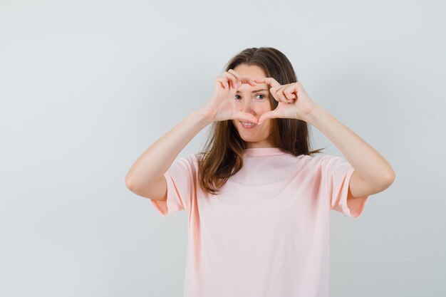 Hembra joven que muestra el gesto del corazón en una camiseta rosa y se ve linda, vista frontal.