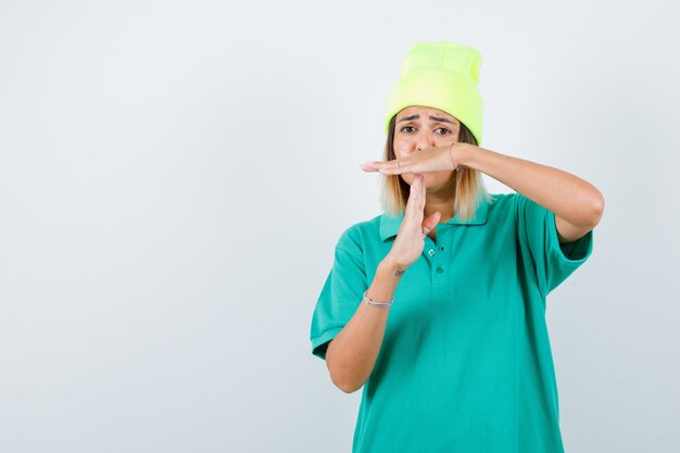 Hembra joven que muestra el gesto de descanso en la camiseta de polo, gorro y mirando molesto, vista frontal.