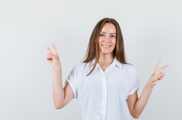 Hembra joven que muestra el gesto del ganador en blusa blanca y parece feliz.