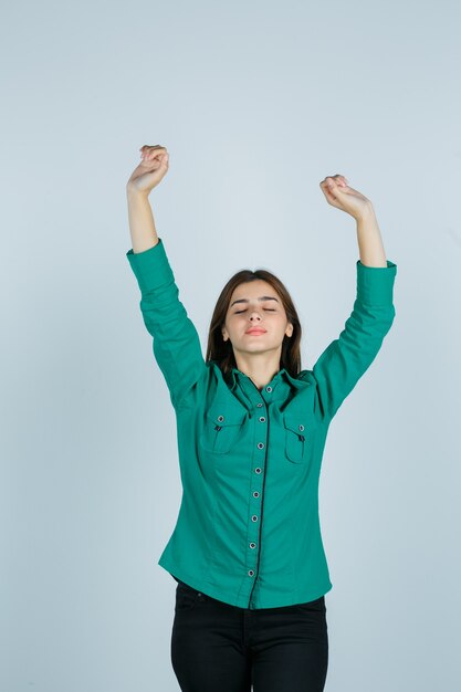 Hembra joven que muestra el gesto del ganador en camisa verde, pantalones y mirando alegre, vista frontal.