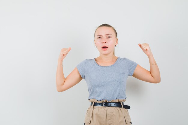 Hembra joven que muestra el gesto del ganador en camiseta, pantalones y que parece afortunado, vista frontal.