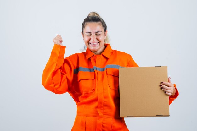 Hembra joven que muestra el gesto del ganador mientras sostiene la caja en uniforme del trabajador y parece jovial. vista frontal.