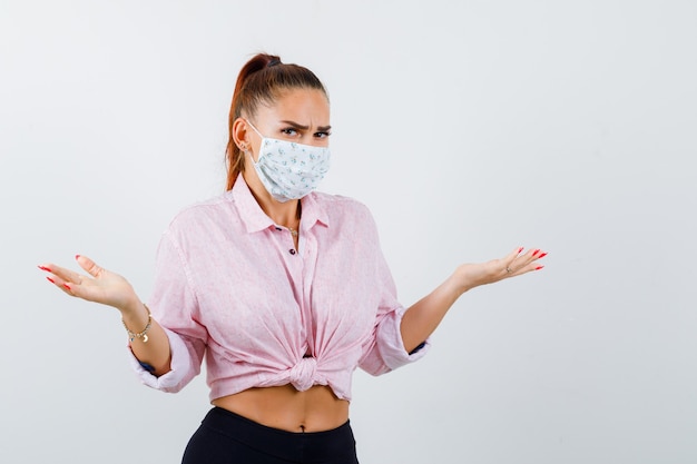 Foto gratuita hembra joven que muestra un gesto de impotencia en camisa, pantalones, máscara médica y parece vacilante. vista frontal.