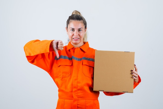 Hembra joven que muestra el pulgar hacia abajo mientras sostiene la caja en uniforme de trabajador y parece disgustado. vista frontal.