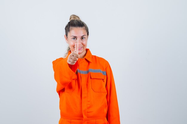 Hembra joven que muestra el pulgar hacia arriba en uniforme de trabajador y parece complacida. vista frontal.