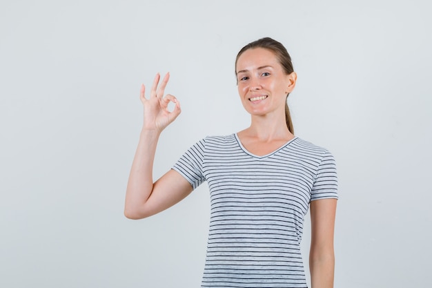 Hembra joven que muestra el signo de ok en camiseta a rayas y parece alegre. vista frontal.