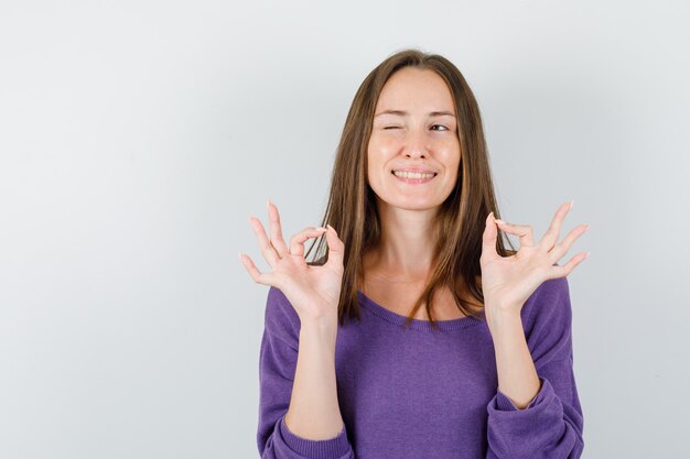 Hembra joven que muestra el signo ok y guiña el ojo en la vista frontal de la camisa violeta.