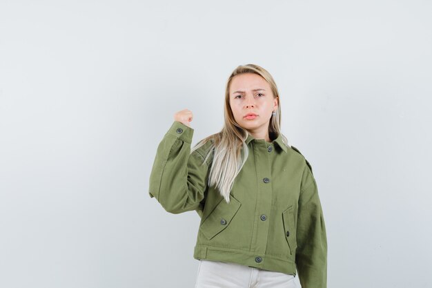 Hembra joven que muestra su poder del brazo en chaqueta verde, jeans y mirando seria, vista frontal.