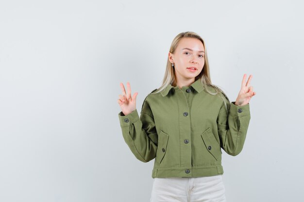 Hembra joven que muestra V-sign en chaqueta verde y mirando alegre, vista frontal.