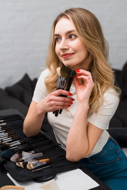 Hembra joven que sostiene algunos pinceles de maquillaje en la mesa