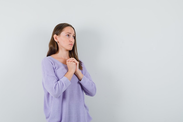 Hembra joven que sostiene las manos combinadas sobre su pecho en una blusa lila y parece tranquila. vista frontal. espacio para texto