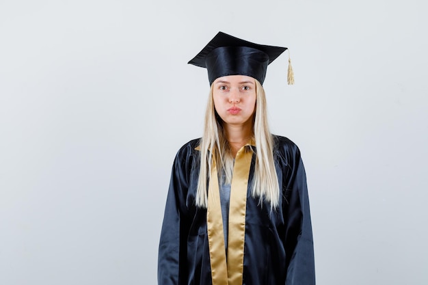 Hembra joven soplando las mejillas mientras mira a la cámara en uniforme de posgrado y parece decepcionado.