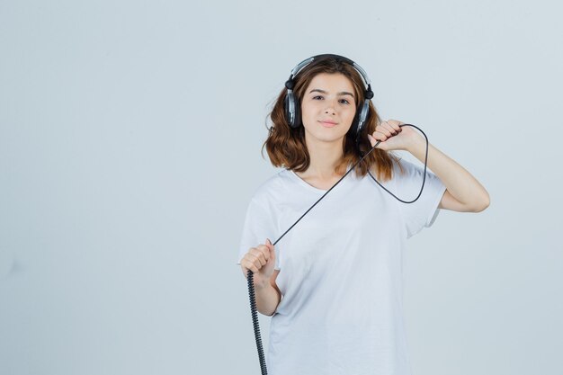 Hembra joven sosteniendo auriculares en camiseta blanca y mirando alegre, vista frontal.