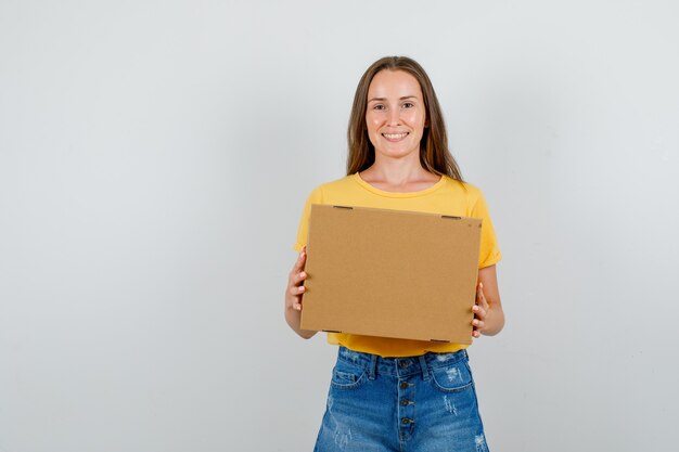 Hembra joven sosteniendo una caja de cartón y sonriendo en camiseta, pantalones cortos