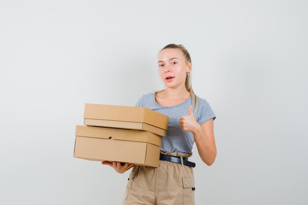 Hembra joven sosteniendo cajas de cartón, mostrando el pulgar hacia arriba en camiseta, pantalones y mirando alegre. vista frontal.