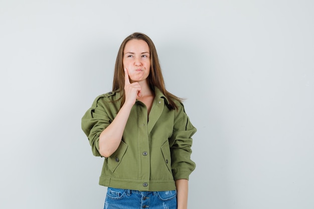 Hembra joven sosteniendo el dedo en la mejilla soplada en chaqueta, pantalones cortos y mirando vacilante, vista frontal.